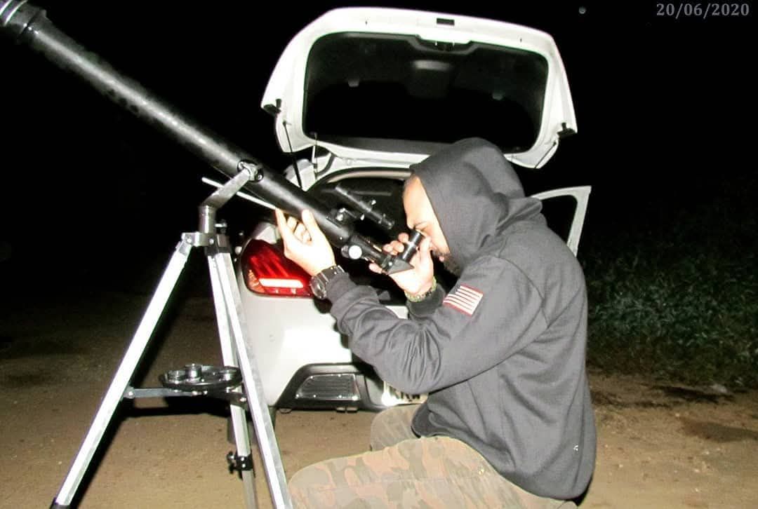 Person in hoodie looking through a large telescope at night, white car in background.